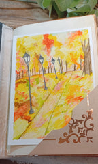 A watercolor painting of an autumn park path with yellow, orange, and red foliage, black lampposts, and trees, displayed on a decorative beige card inside an open book.