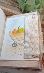 An open vintage-style book or journal page featuring a  watercolor painted autumn scene with a wheelbarrow carrying pumpkins and a yellow squash. A vertical strip of aged book text is layered over the artwork, held in place by a metal clip, with leaves and wood visible around it.