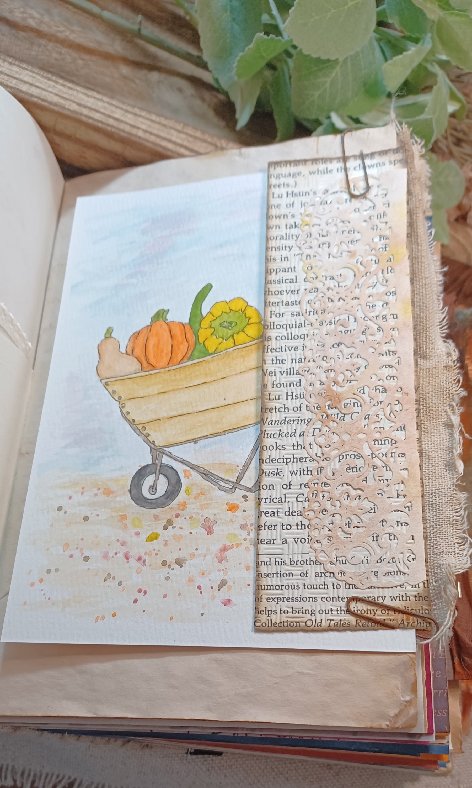 An open vintage-style book or journal page featuring a  watercolor painted autumn scene with a wheelbarrow carrying pumpkins and a yellow squash. A vertical strip of aged book text is layered over the artwork, held in place by a metal clip, with leaves and wood visible around it.