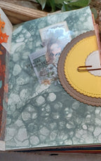 A close-up of a handmade scrapbook or journal page featuring a green and white marble patterned background, layered with a circular brown and yellow paper embellishment and a small photo insert.