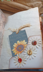 A close-up of a handmade paper journal page featuring a decorative sunflower and layered daisy embellishments, with a blue script tag and rustic wooden background.