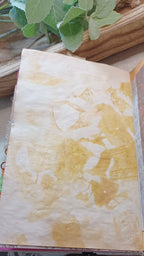 A close-up of a textured, off-white handmade paper sheet with abstract yellow and tan washes from onion peels, resting on a wooden surface with green leaves in the background.