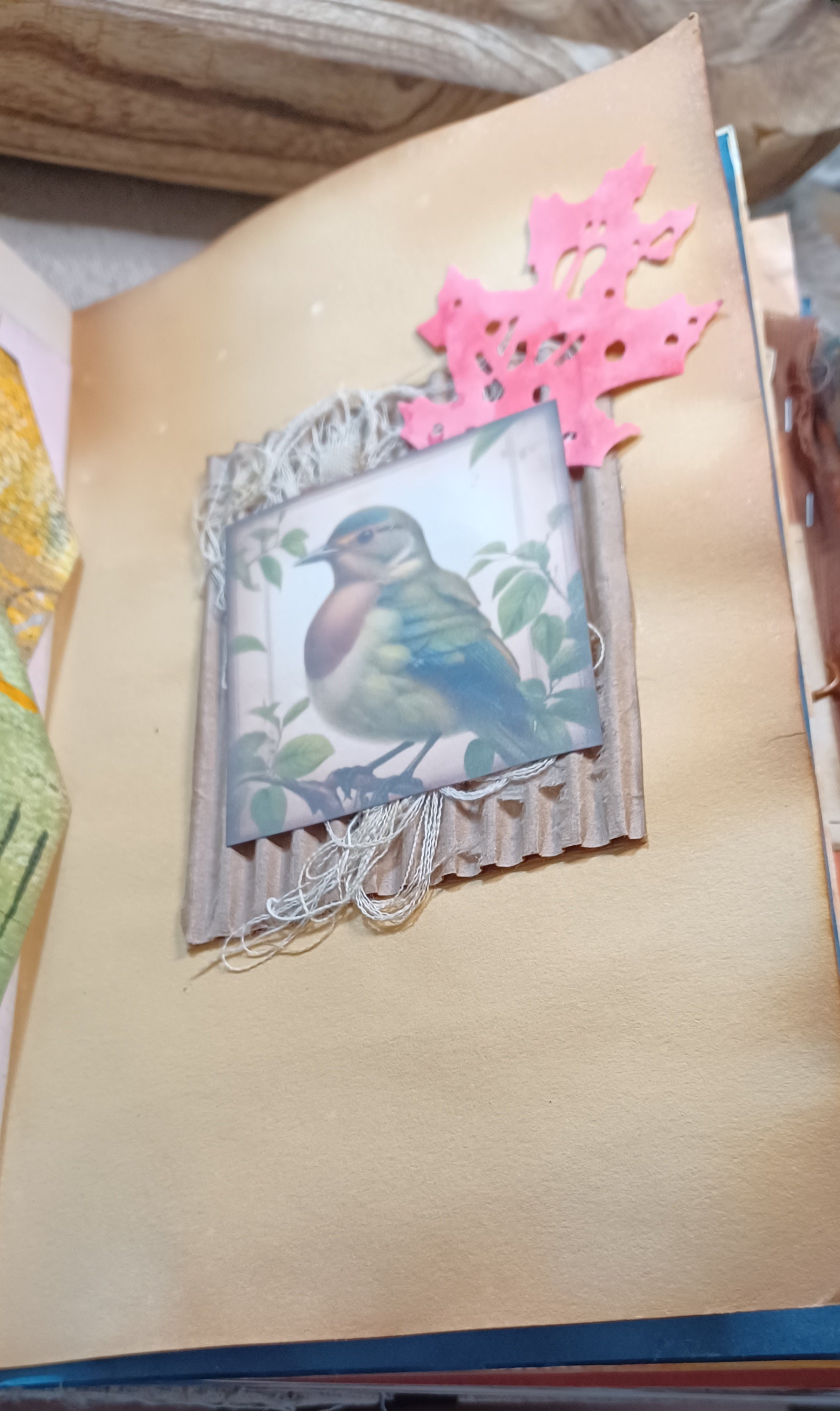 A close-up of a handmade paper collage featuring a vintage-style bird illustration in a rustic corrugated frame, decorated with a orange die-cut and string on a beige background.