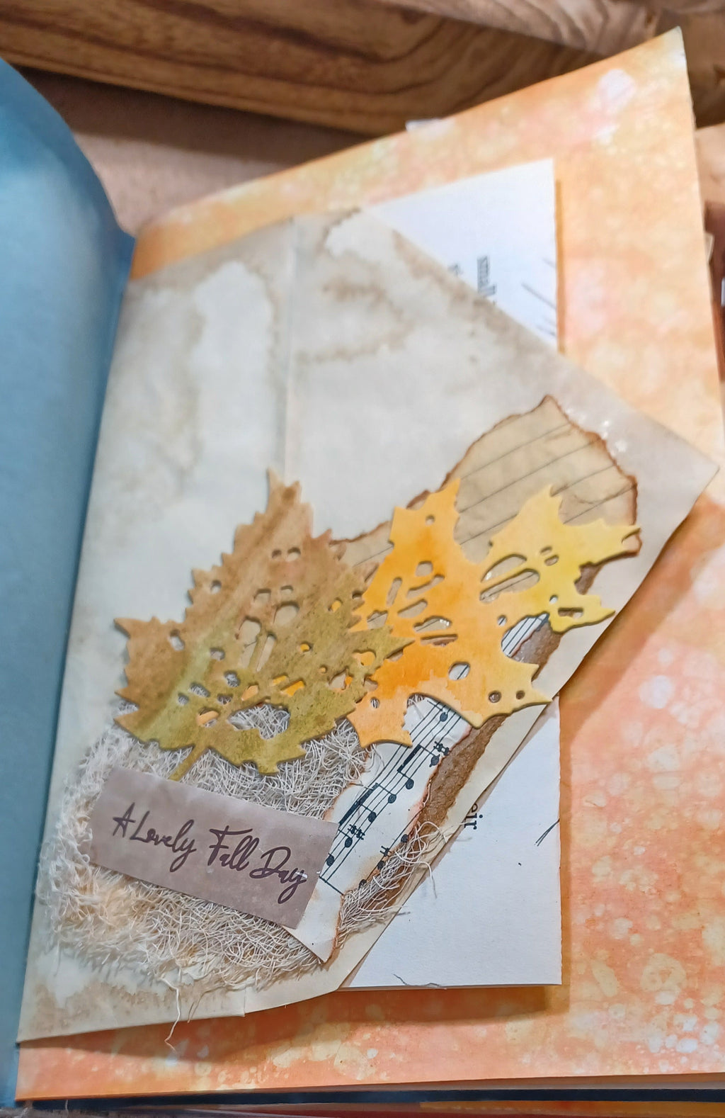A close-up of a handmade layered paper collage featuring autumn leaf shapes in gold and brown, textured beige paper, a piece of paper with musical notes, and a small tab reading 'A Lovely Fall Day'.
