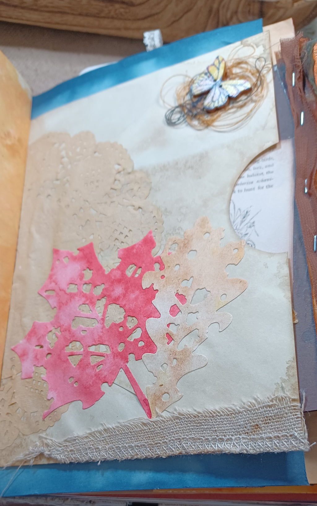 A close-up of a handmade mixed-media paper collage featuring layered beige textured papers, a orange watercolor maple leaf, a tan leaf cutout, a small butterfly embellishment, and a piece of fabric along the bottom edge.