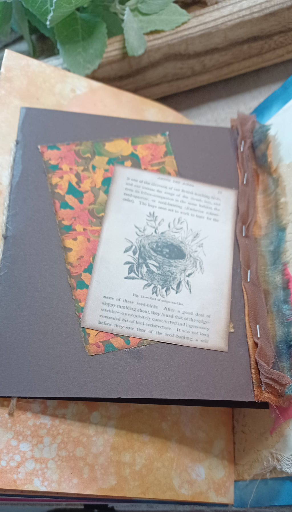 A close-up of a handmade journal page featuring a dark brown cover, a patterned autumn leaf insert, a faux vintage paper page with an illustration of a bird's nest, and rustic fabric tabs along the side.