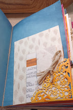 A close-up of a handmade paper journal or scrapbook page with a green page, patterned insert, folded white paper, a small orange label, burlap ribbon, and a yellow die-cut decorative overlay.