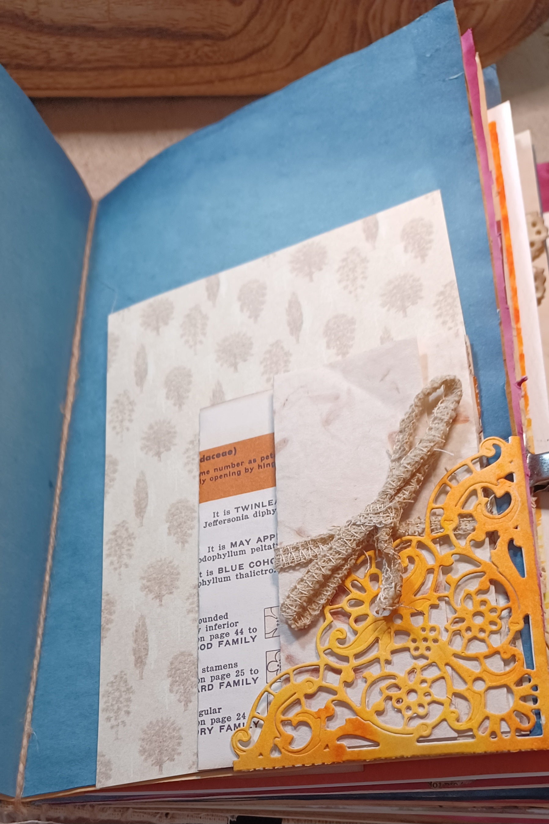 A close-up of a handmade paper journal or scrapbook page with a green page, patterned insert, folded white paper, a small orange label, burlap ribbon, and a yellow die-cut decorative overlay.