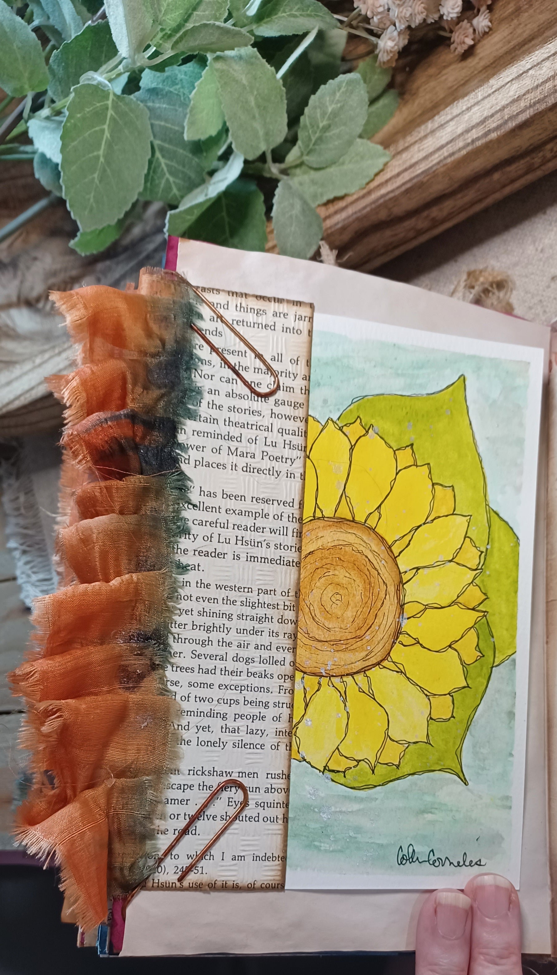 A close-up of a handmade journal page featuring a yellow and green sunflower watercolor painting, layered over a book page with a textured orange ribbon bookmark. A finger is visible holding the page, with decorative green leaves and a wooden background in the scene.