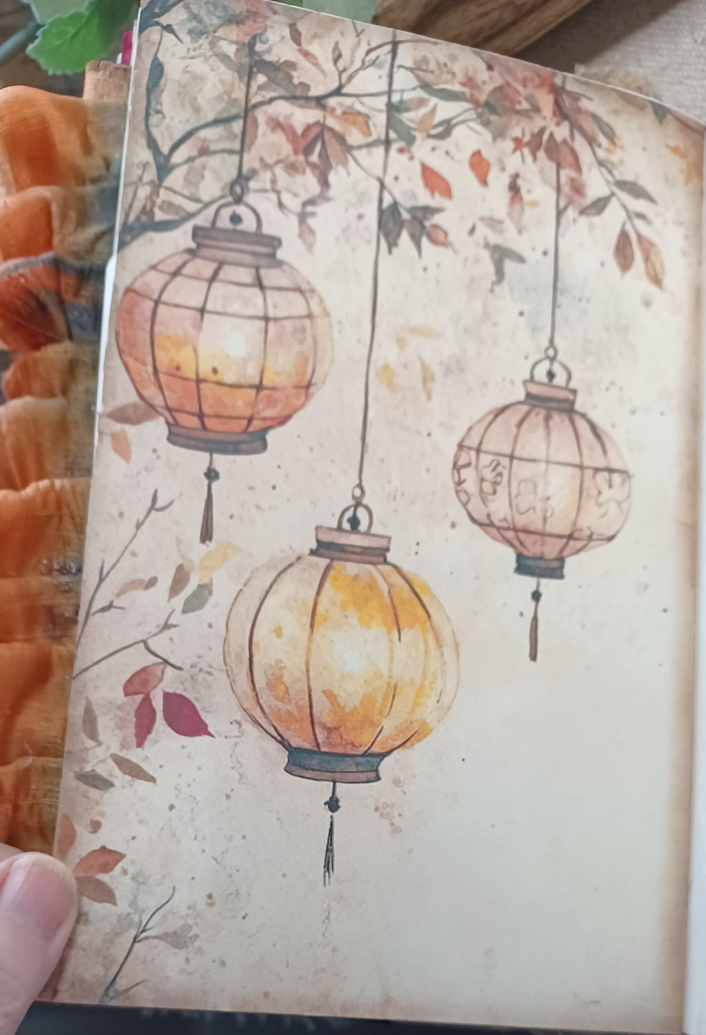 A close-up of a printed card or notebook page featuring an illustration of three hanging lanterns with autumn leaves on a warm beige background. A hand is partially visible holding the page, and orange fabric appears along the left edge.