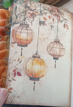 A close-up of a printed card or notebook page featuring an illustration of three hanging lanterns with autumn leaves on a warm beige background. A hand is partially visible holding the page, and orange fabric appears along the left edge.