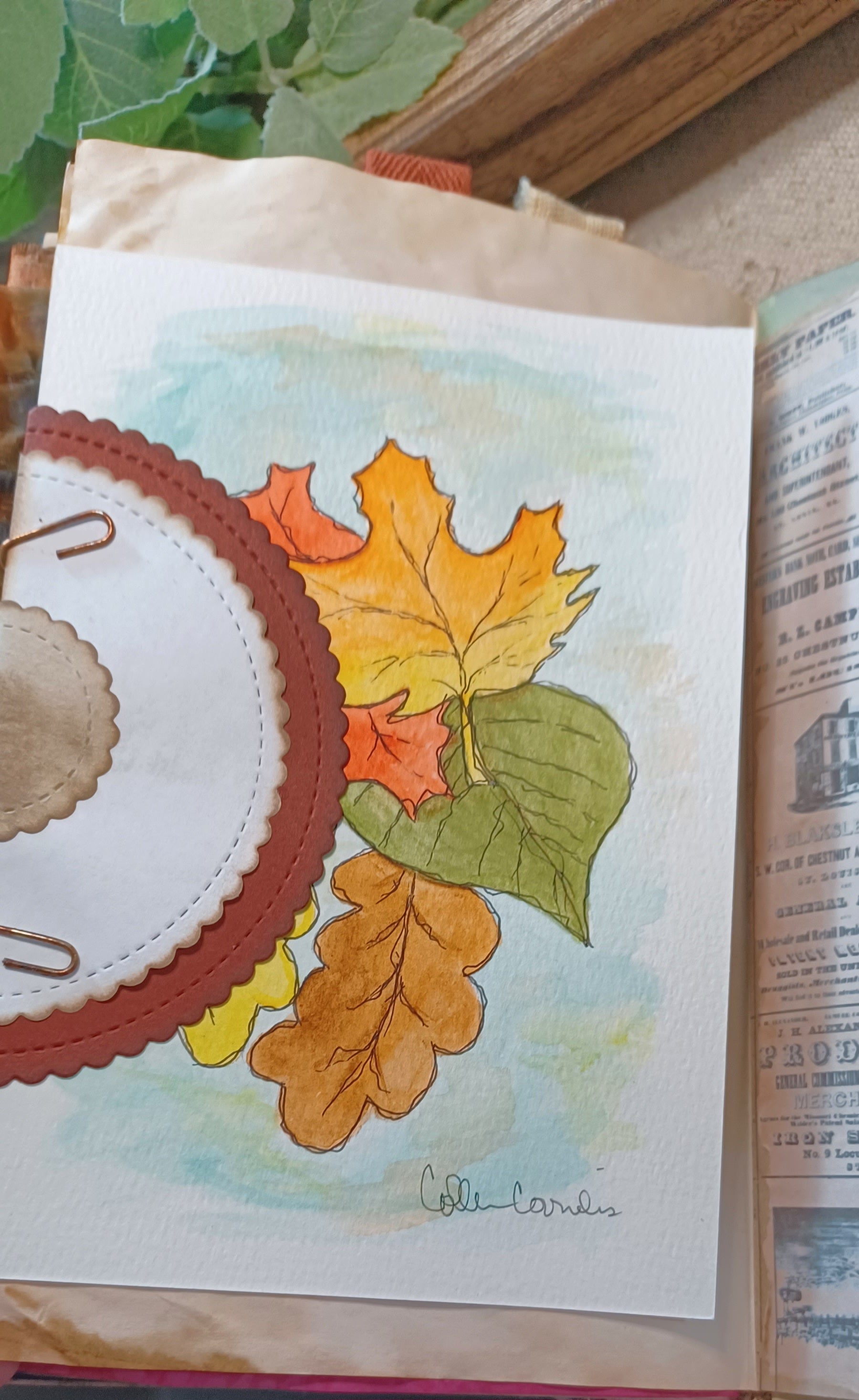 A close-up of a handmade paper craft or scrapbook page featuring a watercolor painting of autumn leaves in orange, yellow, and green on a white background, layered over a coffee dyed paper sheet and a vintage book page.