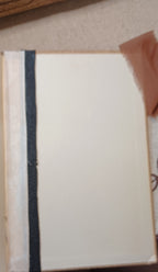 A close-up of a blank off-white notebook or journal cover with a dark navy blue vertical stripe down the left side, resting on a textured beige surface.