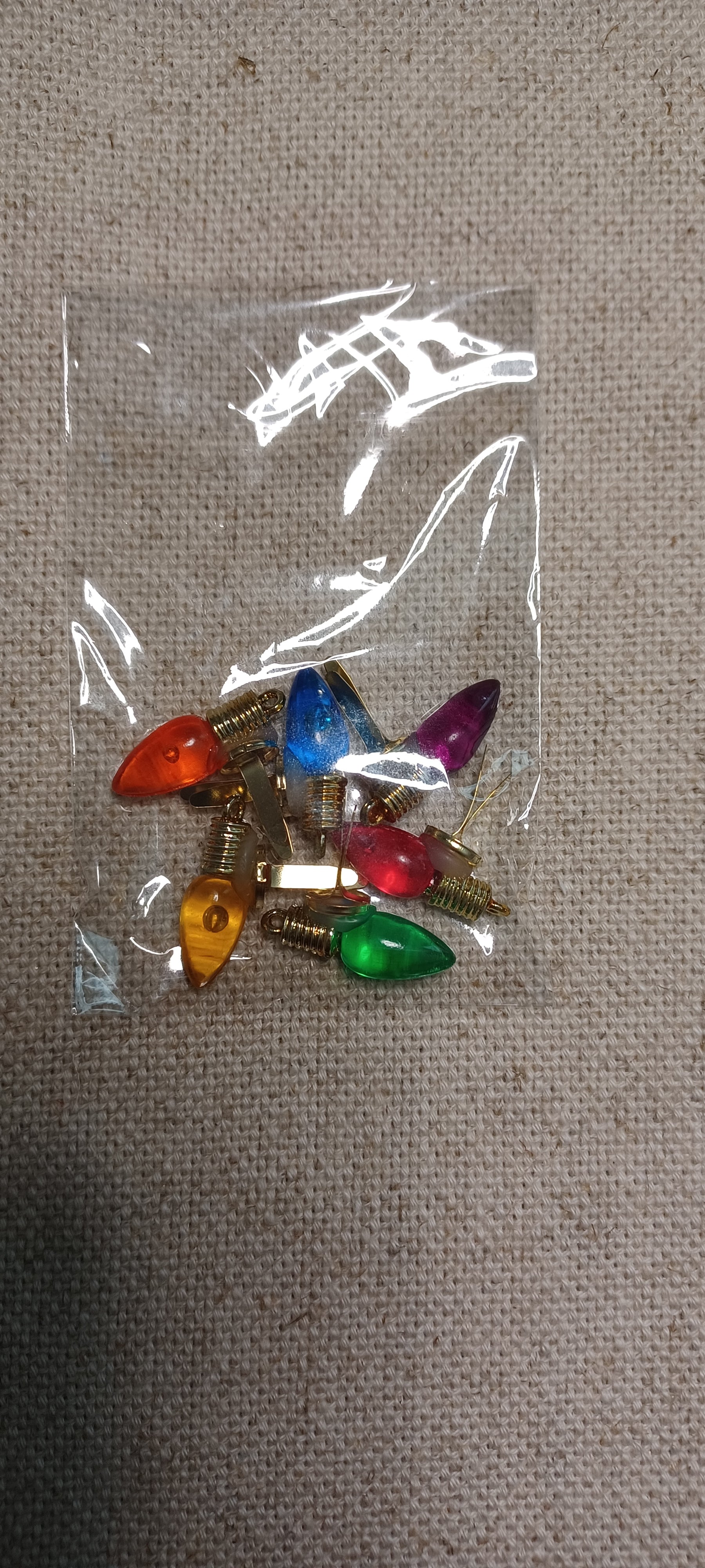 A small clear plastic bag containing several colorful Christmas light bulbs with gold brad base , placed on a beige fabric background.