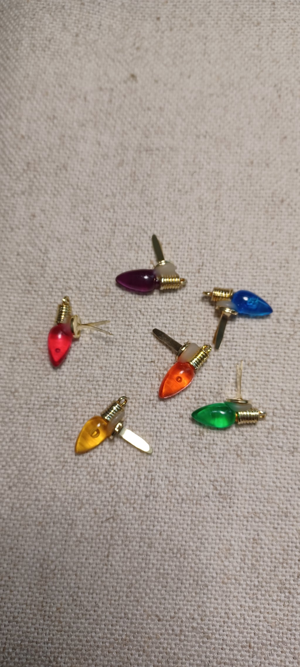 A set of seven Christmas light-shaped paper fastener with gold brad base in red, purple, blue, orange, green, yellow, and white, arranged on a beige textured fabric background.