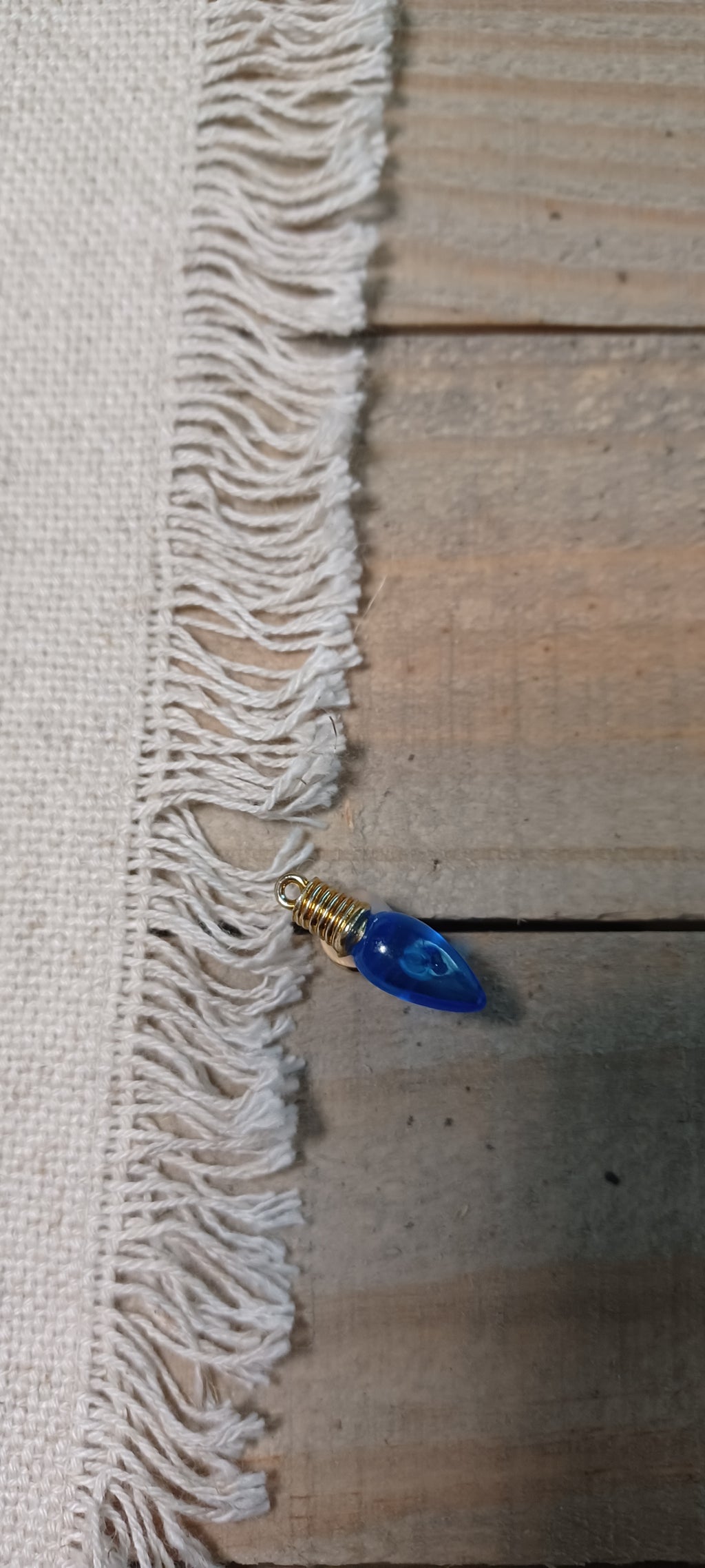A close-up of a white woven fabric with fringed edges laid on a rustic wooden surface, with a small blue teardrop-shaped Christmas light paper fastener with gold brad base resting near the fringe.
