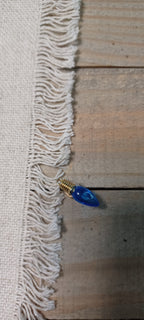 A close-up of a white woven fabric with fringed edges laid on a rustic wooden surface, with a small blue teardrop-shaped Christmas light paper fastener with gold brad base resting near the fringe.
