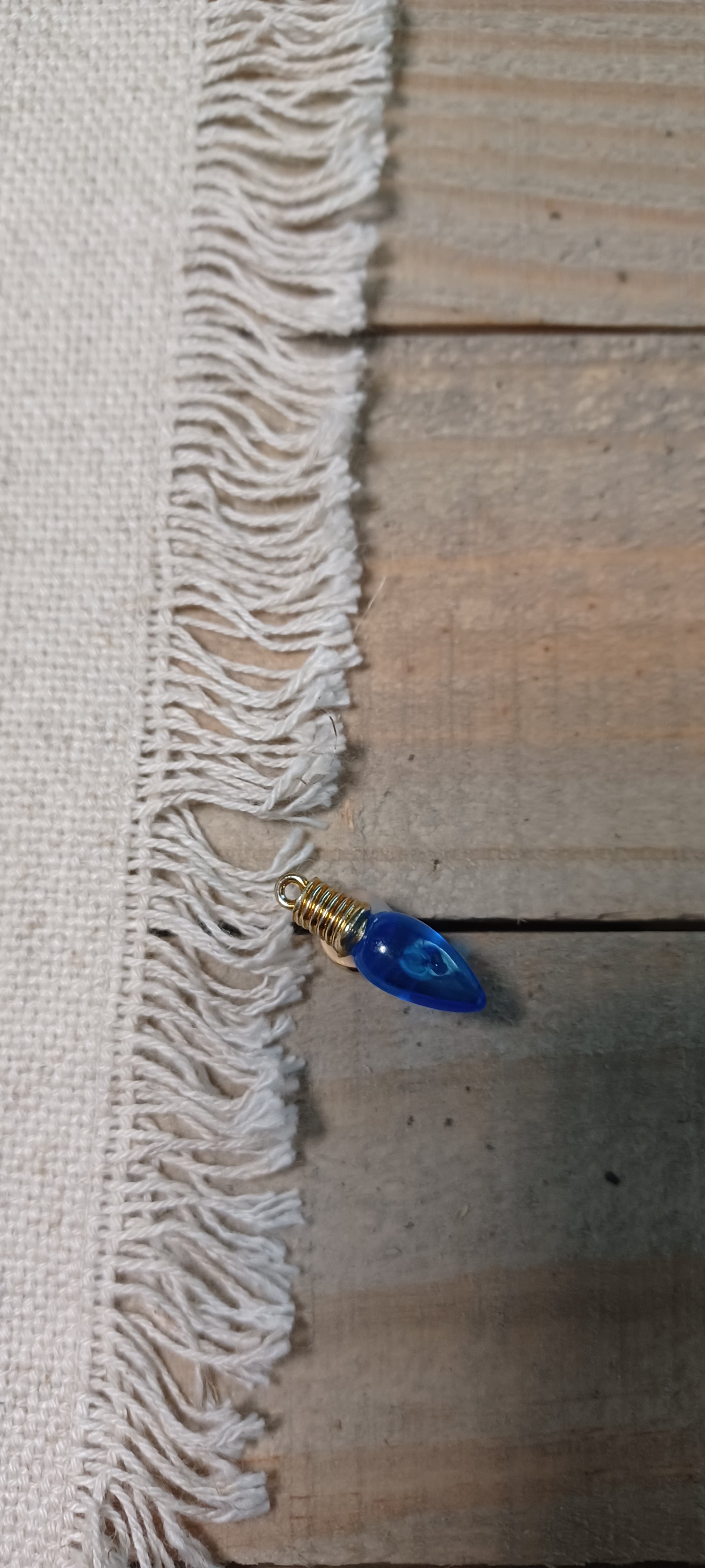 A close-up of a white woven fabric with fringed edges laid on a rustic wooden surface, with a small blue teardrop-shaped Christmas light paper fastener with gold brad base resting near the fringe.