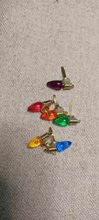 Six small Christmas light-shaped paper fasteners with gold-tone metal bases and colorful plastic bulbs in red, orange, green, yellow, blue, and purple, arranged on a beige textured fabric background.
