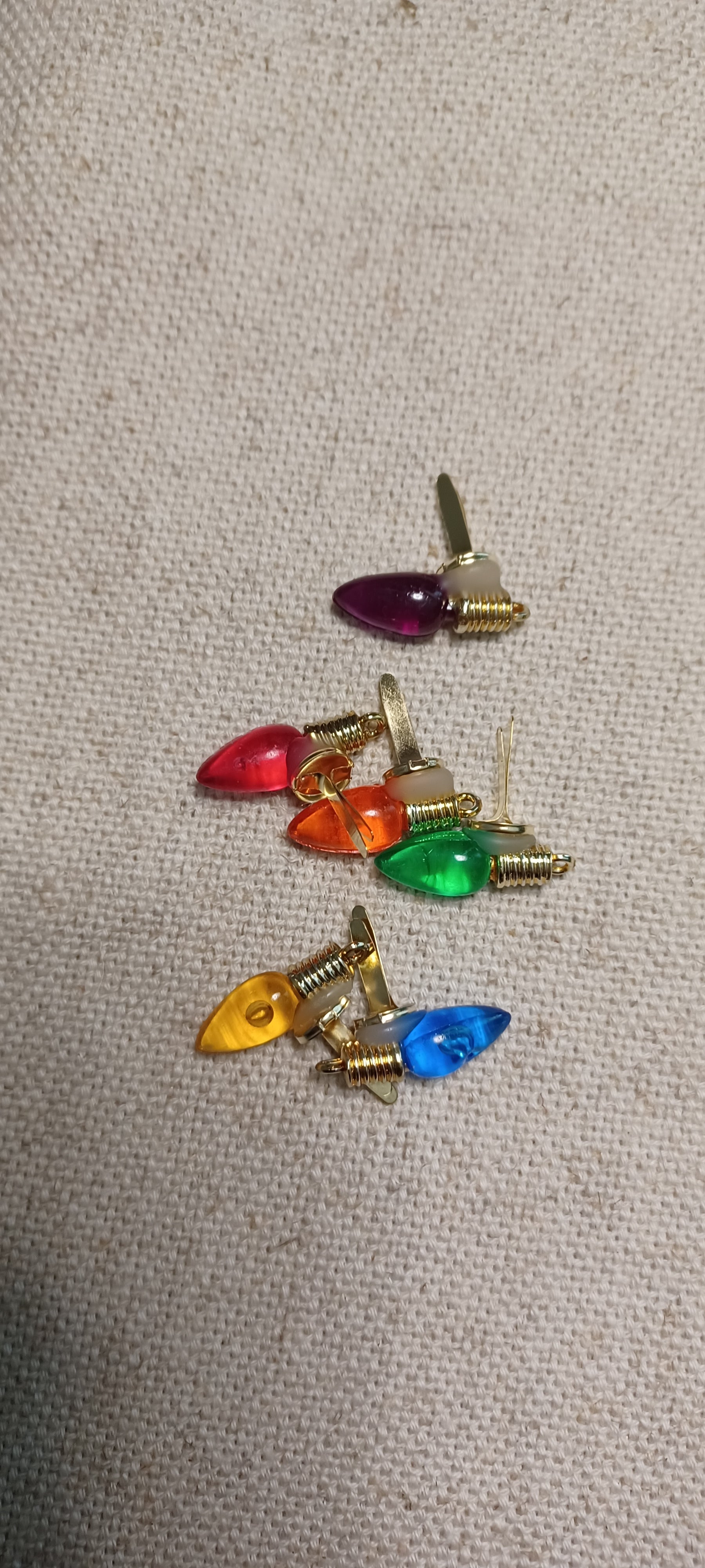 Six small Christmas light-shaped paper fasteners with gold-tone metal bases and colorful plastic bulbs in red, orange, green, yellow, blue, and purple, arranged on a beige textured fabric background.