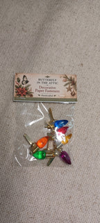 A small clear plastic bag containing decorative paper fasteners with colorful butterfly and floral designs, attached to a printed header card that reads 'Butterfly in the Attic Decorative Paper Fasteners Handmade by J.' The product is photographed on a light beige fabric background.