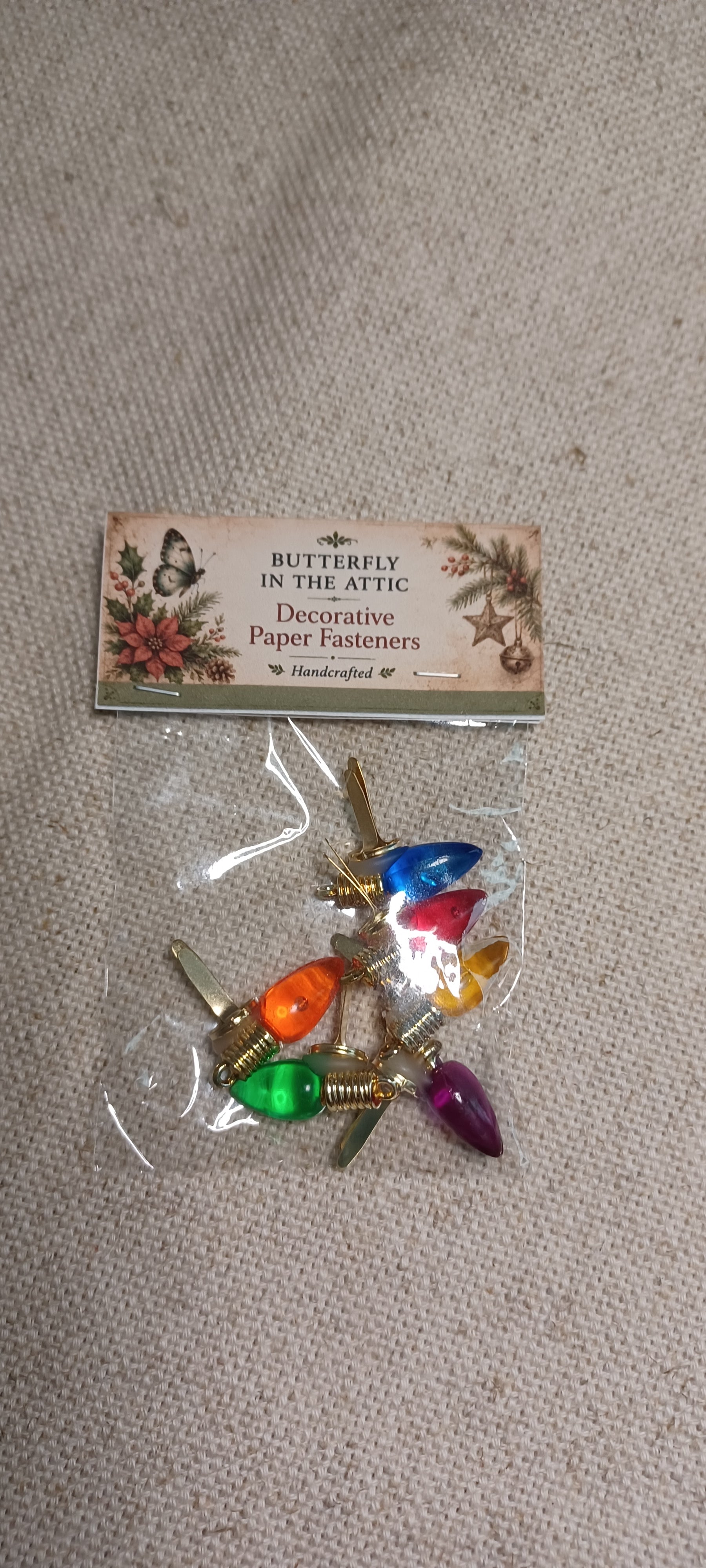 A small clear plastic bag containing decorative paper fasteners with colorful butterfly and floral designs, attached to a printed header card that reads 'Butterfly in the Attic Decorative Paper Fasteners Handmade by J.' The product is photographed on a light beige fabric background.