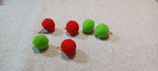 Six small felt pom-pom decorative paper fasteners in alternating red and green on metal brads, arranged on a textured beige fabric background.