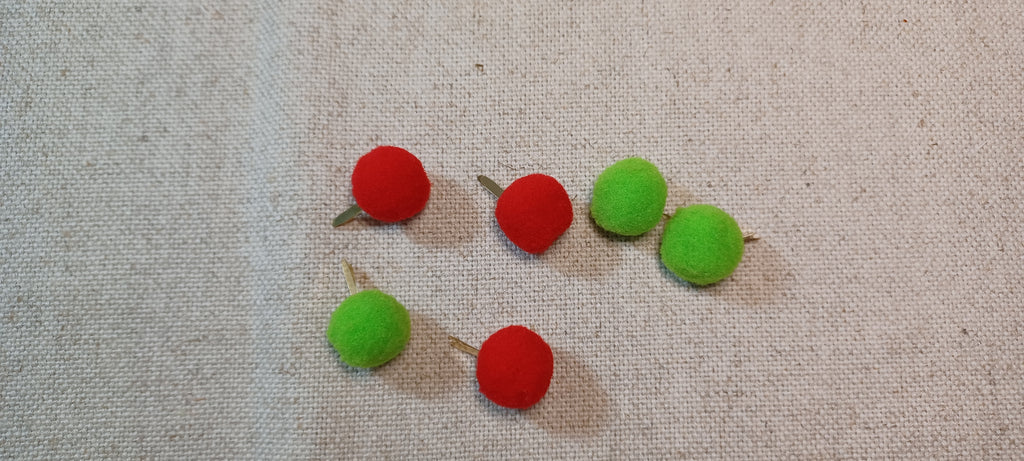six small felt pom-pom decorative paper fasteners , alternating red and green, arranged on a textured beige linen fabric background.