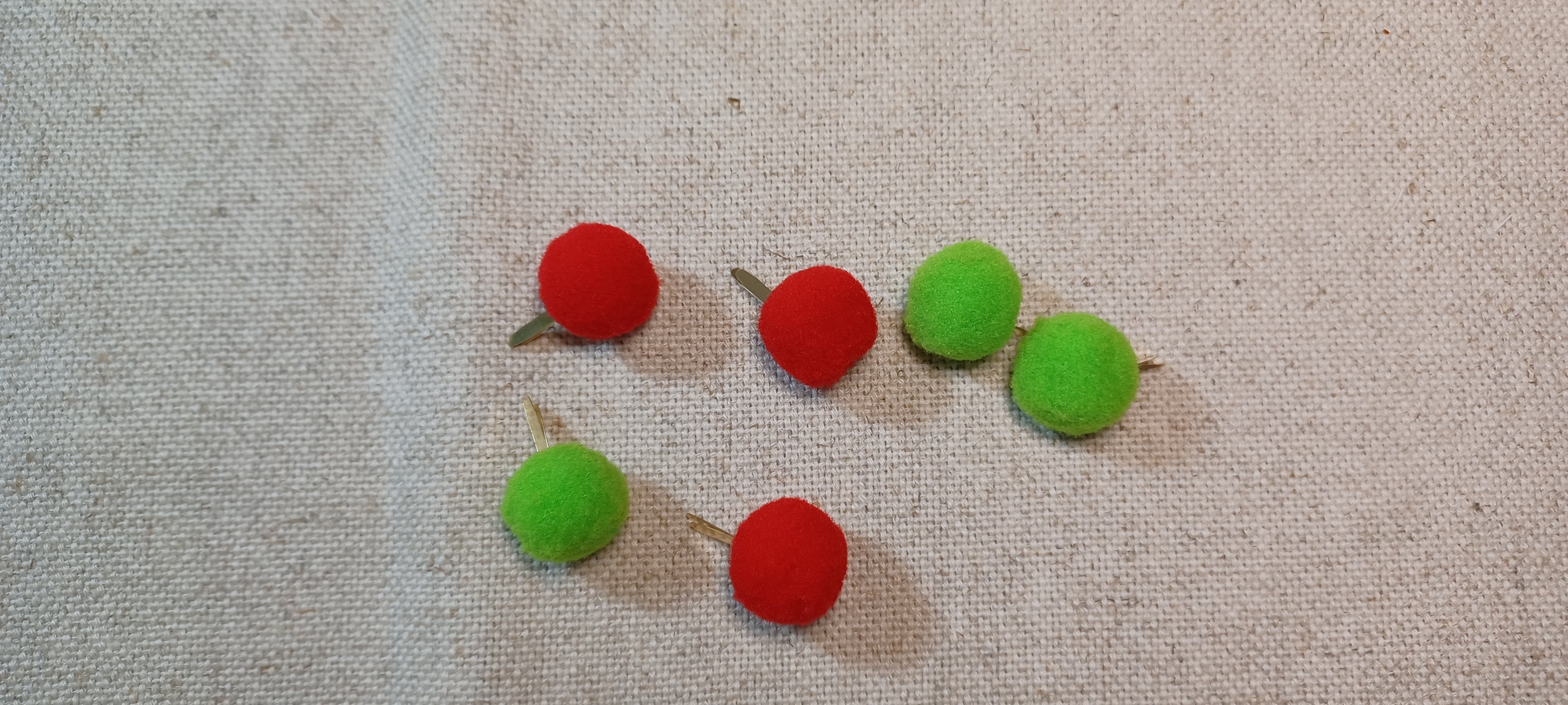 six small felt pom-pom decorative paper fasteners , alternating red and green, arranged on a textured beige linen fabric background.