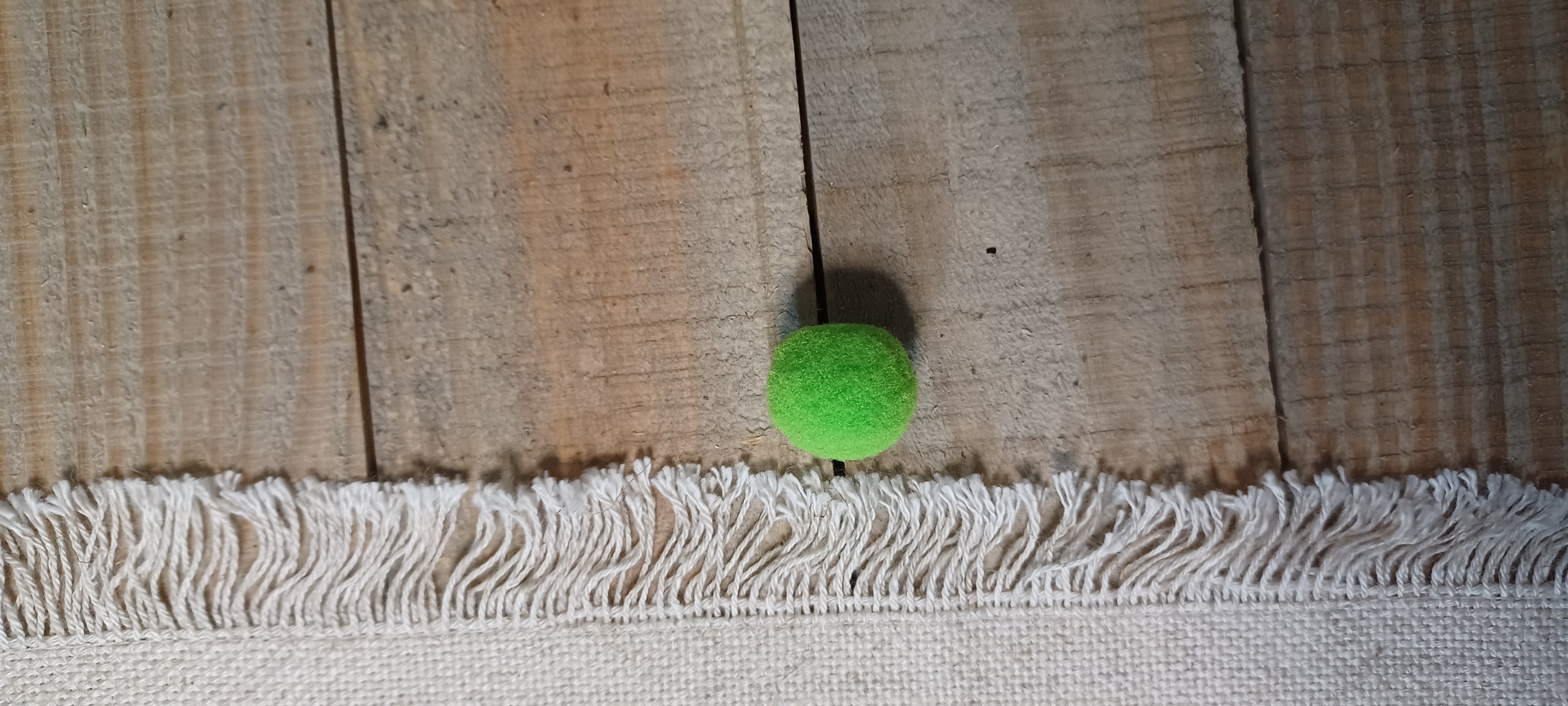 A small bright green pom pom ball decorative paper fasteners  sits on a rustic wooden plank floor with a white fringed textile runner in the foreground.