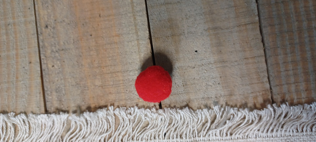 A small red pom-pom ball decorative paper fastener from view above , positioned above a white fringed textile on a rustic wooden plank surface.