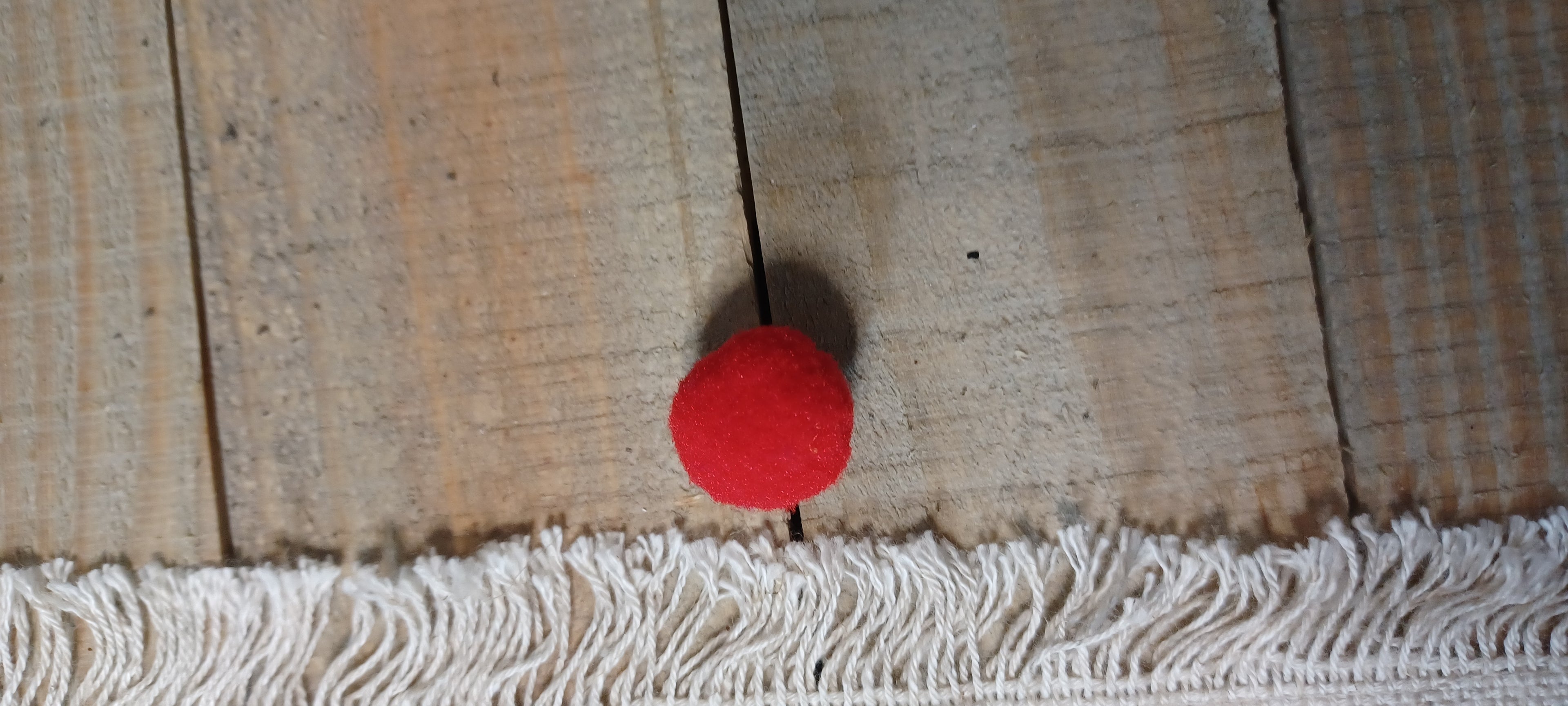 A small red pom-pom ball Paper Fastener, positioned above a white fringed textile on a rustic wooden plank surface.