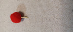 A small red pom-pom decorative paper fasteners  with a gold metal brad back, resting on a textured beige linen fabric background.