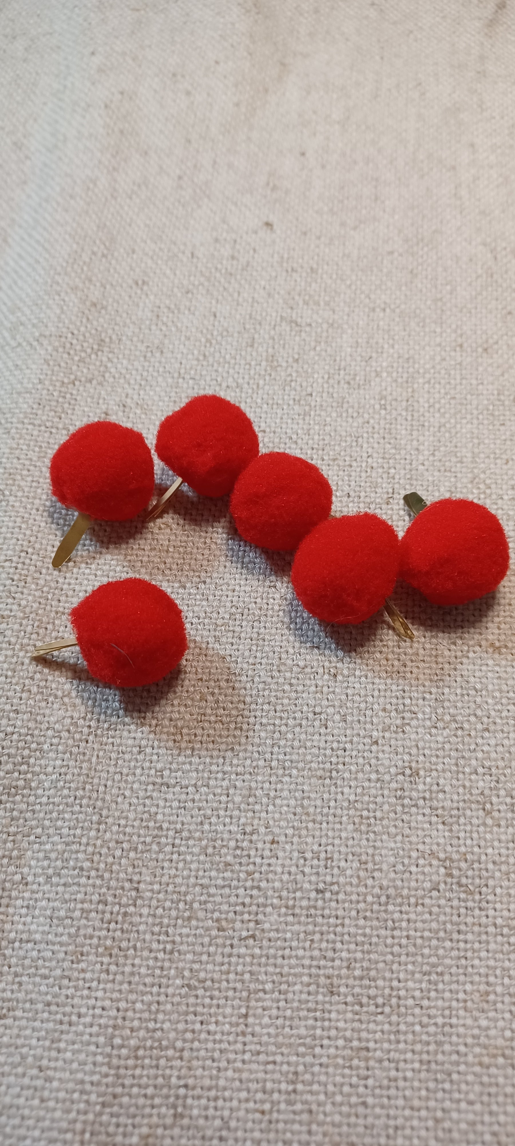 Five small red pom-pom Paper Fastener with gold-tone wire stems, arranged on a textured beige fabric background.