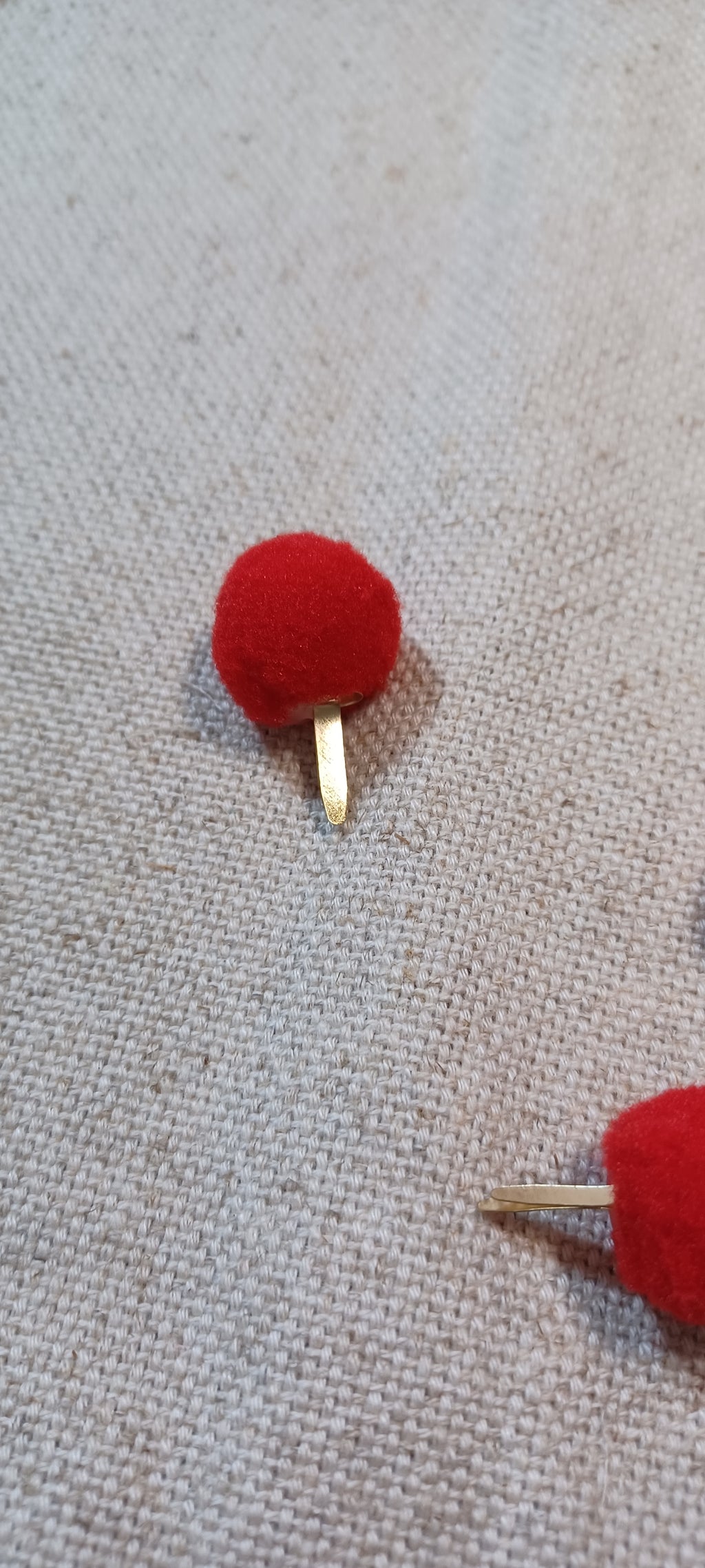 A close-up of a small red pom-pom Paper Fastener with a gold-tone metal post, placed on a textured beige fabric background.