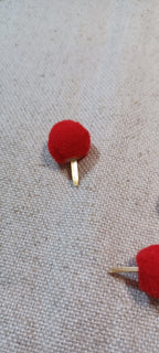 A close-up of a small red pom-pom Paper Fastener with a gold-tone metal post, placed on a textured beige fabric background.