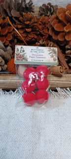 A small clear plastic bag containing six bright red felt pom-poms, with a printed paper header card reading 'Butterfly in the Attic Decorative Paper Fasteners.' The package is photographed on a light fabric surface with pinecones and warm decorative props in the background.