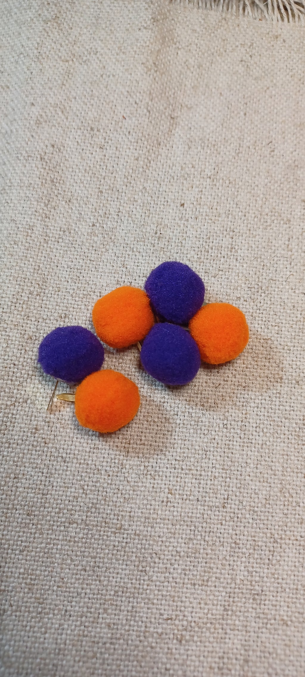 Five small felt pom-poms in purple and orange paper fasteners arranged on a beige woven fabric background.