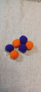 Five small felt pom-poms in purple and orange paper fasteners arranged on a beige woven fabric background.