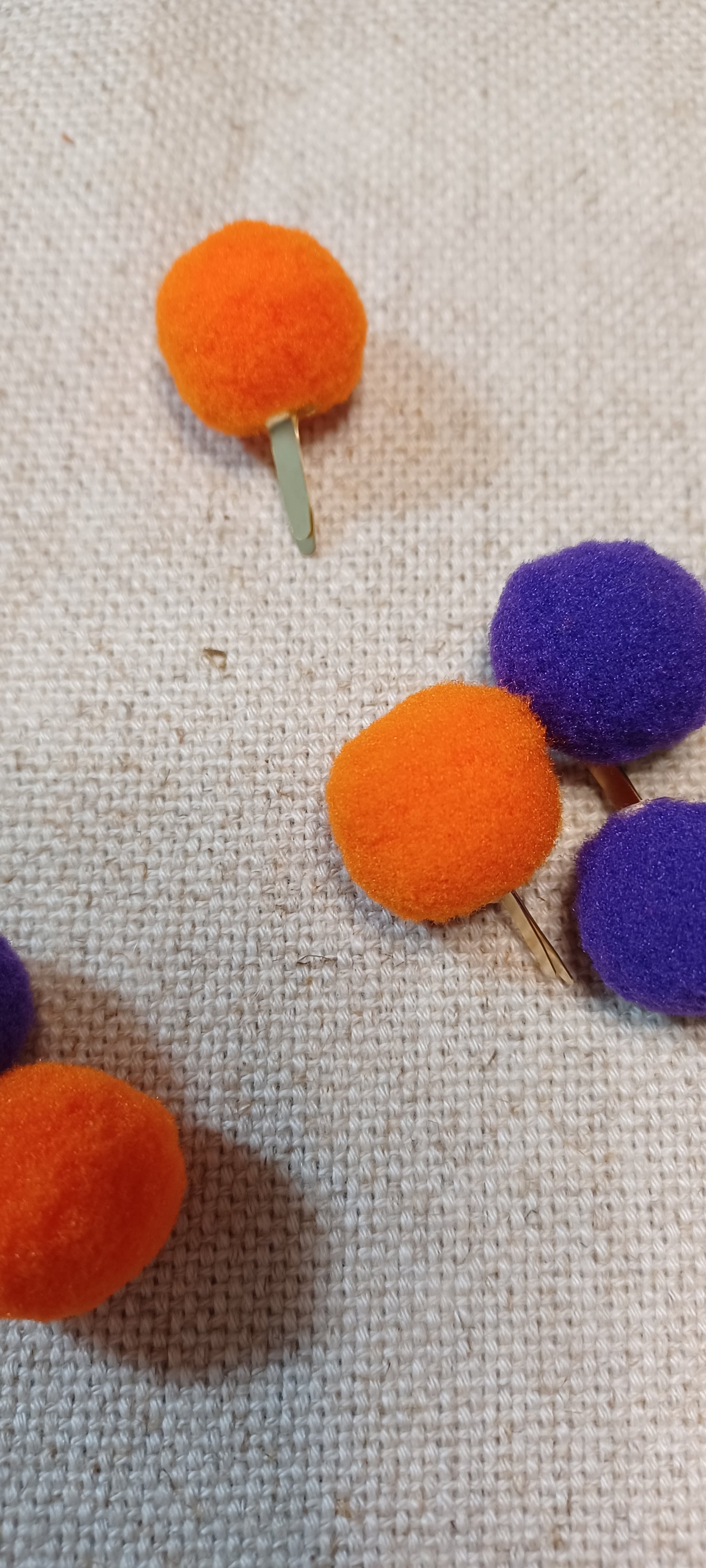 Small felt pom-pom paper fasteners in orange and purple attached to thin metal brads, arranged on a textured beige fabric background.
