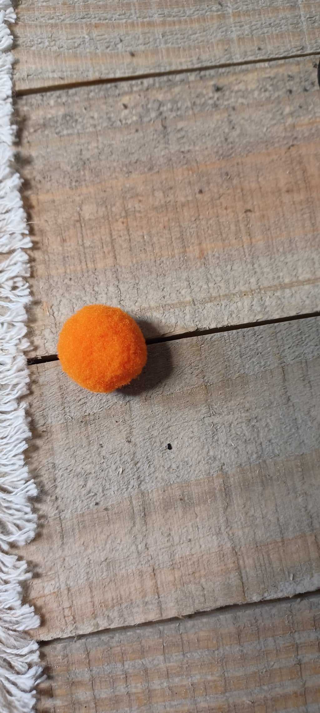 A small orange pom-pom ball  paper fastener resting on a rustic wooden plank surface next to a white fringed textile.