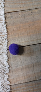 A small purple felt pom-pom ball paper fastener resting on a rustic wooden plank surface next to a white fringed textile.