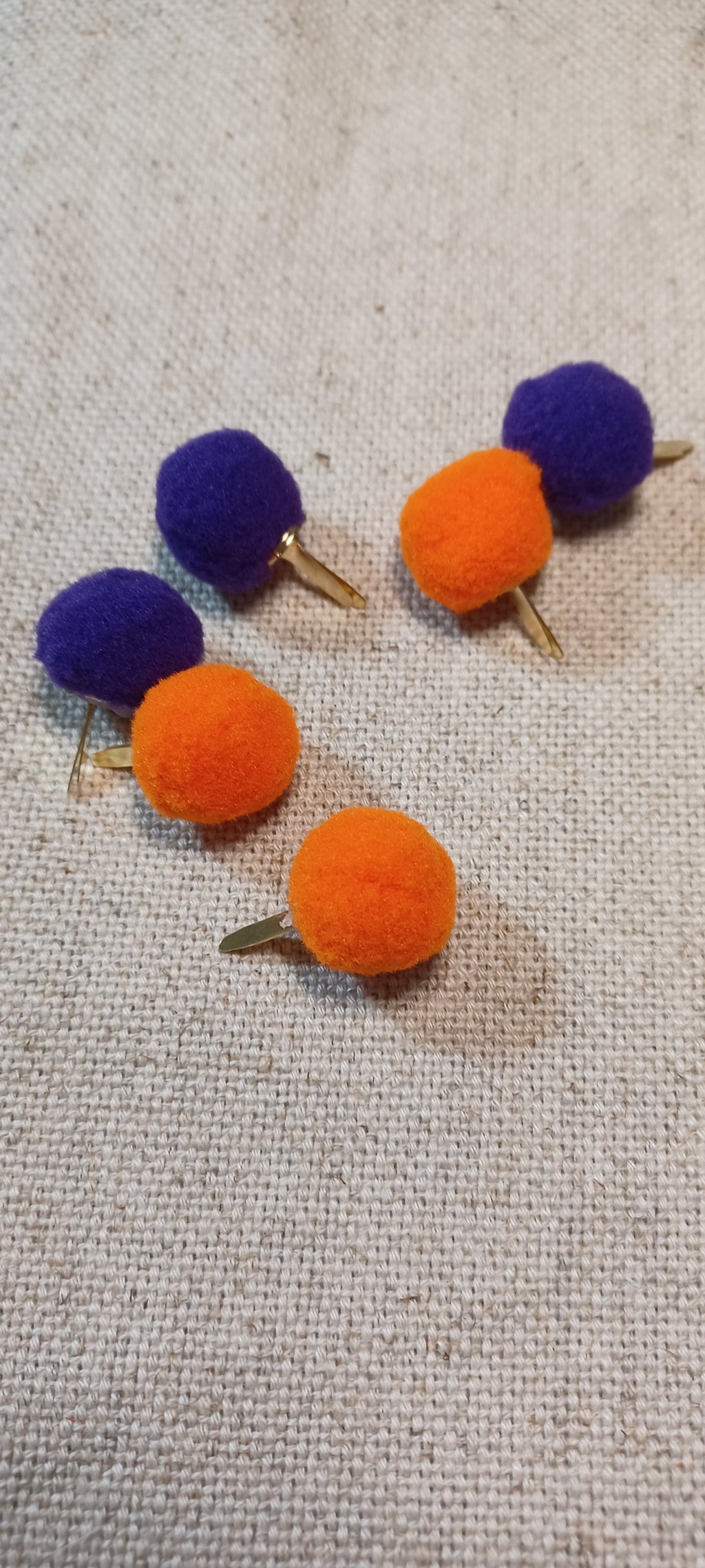 Five small pom-pom paper fastener on a textured beige fabric background. Three pom-poms are bright orange with gold-tone brads , and two are deep purple with gold-tone brads