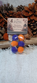 A small clear plastic bag containing six decorative paper fasteners in Halloween colors, purple and orange, attached to a printed card that reads 'Butterfly in the Attic Decorative Paper Fasteners.' The package is styled with pinecones and a fringed fabric surface.