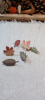 Six small leaf-shaped accessories in autumn colors are arranged on a textured off-white fabric surface, with pinecones and dried foliage in the background.