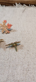 A close-up of small autumn-themed metal paper fasteners resting on a textured beige linen fabric. The paper fasteners include a maple leaf design in orange and gold, and a pine tree design in gold and green.