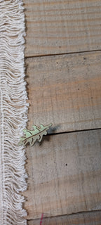 A small metallic leaf-shaped paper fastener lies on a rustic wooden surface next to a white fringed textile.