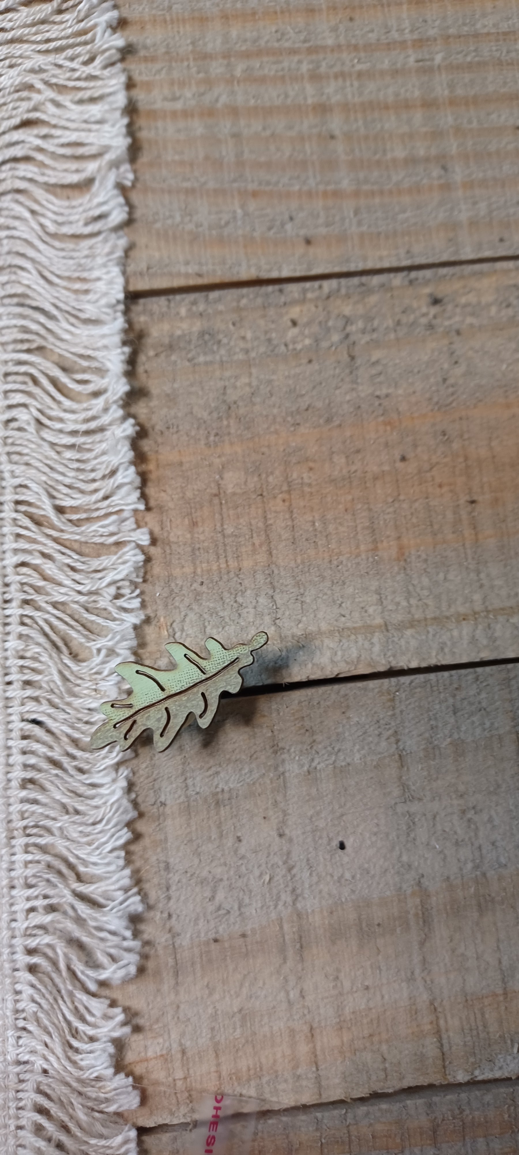 A small metallic leaf-shaped paper fastener lies on a rustic wooden surface next to a white fringed textile.