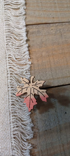 A small wooden leaf-shaped paper fastener with a tan and orange gradient, resting on a rustic wooden surface next to a white fringed textile.