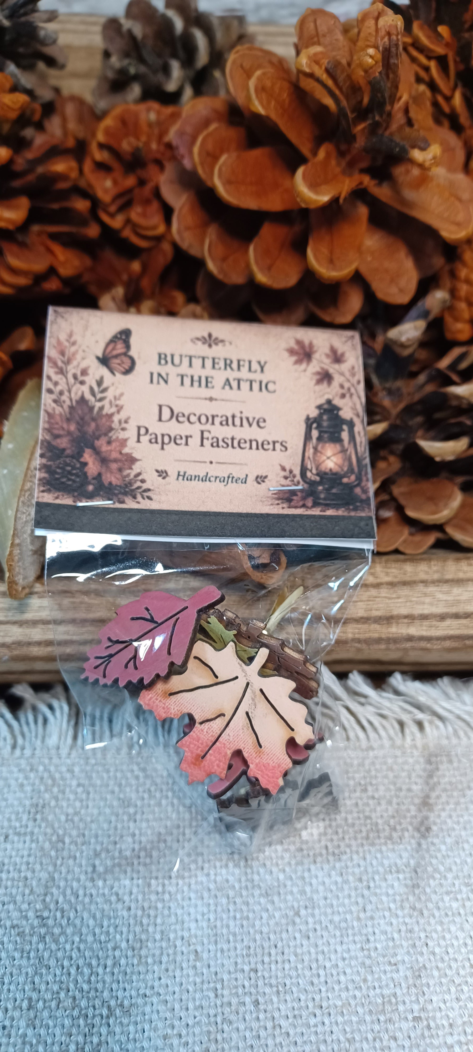 A small clear plastic bag containing Fall Leaf-themed decorative paper fasteners, placed in a bag with a topper where the front of a printed card that reads 'Butterfly in the Attic Decorative Paper Fasteners Handcrafted.' The product sits on a light fabric surface with pinecones and wood in the background.